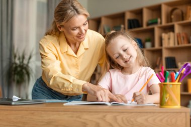 Caring mother helping to adorable little daughter with homework, sitting at table in living room, smart girl doing school tasks, studying, homeschooling