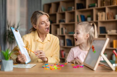 Speech training for kids. Professional female specialist training with little girl at cabinet, teaching her right articulation exercises, showing something on clipboard