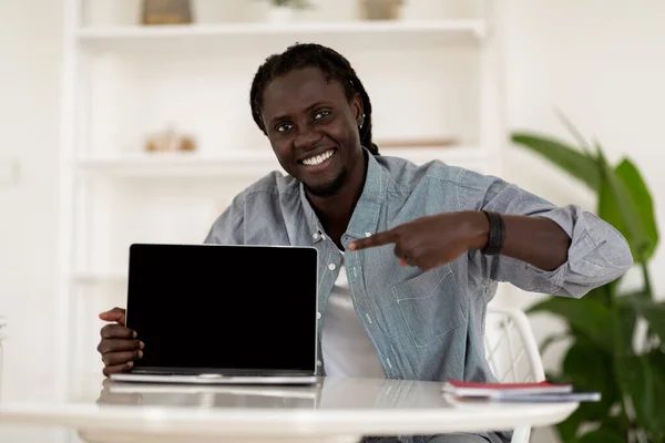 Happy Handsome Black Man Sitting At Desk And Pointing At Blank Laptop Screen At Home Office, Smiling African American Male Recommending New Website Or Online Offer, Mockup Image With Copy Space