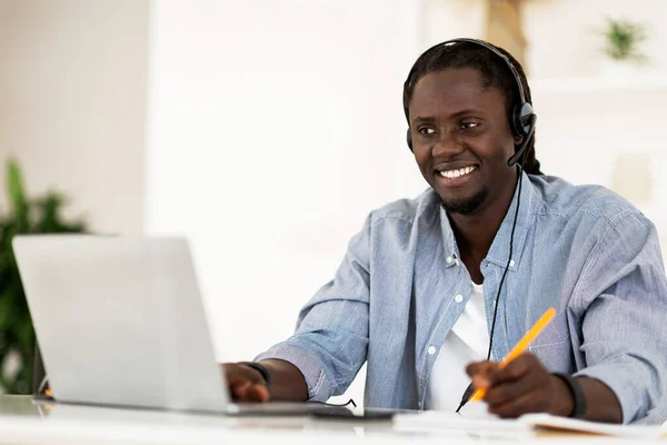 Online Education. Black Smiling Man In Headset Study With Laptop At Home, Handsome African American Man Sitting At Desk, Looking At Computer Screen And Taking Notes, Enjoying Distance Learning