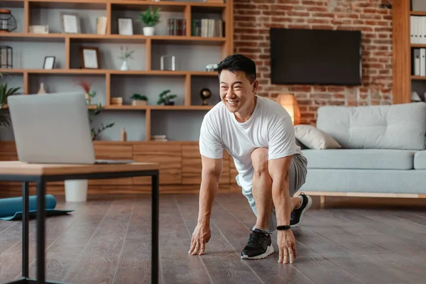 Home training. Happy asian mature man doing stretching exercises for legs in front of laptop, watching online tutorials and smiling, enjoying domestic workout