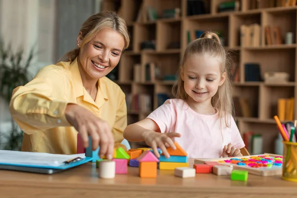 Games and education concept. Positive professional female teacher exercising with little smart girl, playing wooden development blocks at classroom, sitting at desk