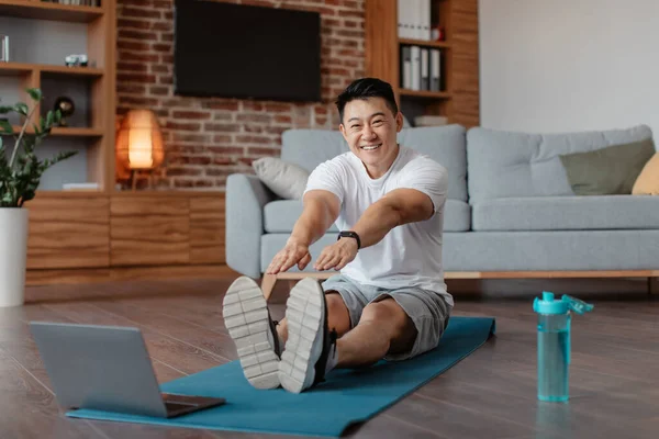 Home training concept. Positive sporty korean man stretching leg muscles in front of laptop, watching video tutorials online, enjoying healthy lifestyle, copy space