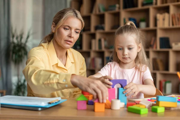 Liltle preschooler girl playing with wood blocks and teacher educador help, sitting at table in office. Homeshooling, learning community, montessori school