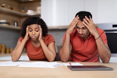 Upset despaired millennial african american wife and husband in same t-shirts think and work with documents in kitchen interior. Problems with paying taxes, loan, mortgage and debts, home bookkeeping