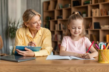 Happy female tutor sitting at table with smart little girl, helping child with homework, preparing school assignments together at home. Homeschooling concept.