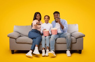 Joyful black family of three sitting on sofa and watching movie, eating popcorn from bucket laughing at show, woman holding remote control isolated on yellow studio wall