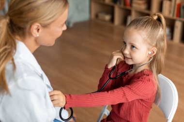 Pretty little girl visiting pediatrician and playing with female doctor, listening docs heart beating with stethoscope, sitting in clinic and smiling