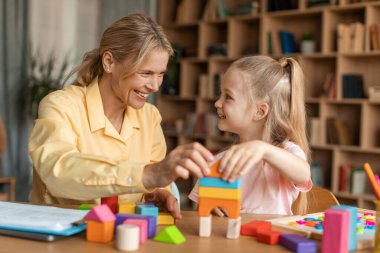 Cheerful child development specialist having session with cute little girl, sitting at desk and playing with colorful wooden bricks, looking and smiling at each other