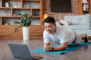Positive asian mature man standing in elbow plank position, training at home and watching online sports video on laptop. Male exercising abs muscles, doing domestic workout
