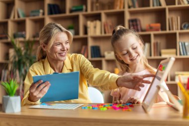 Pretty little girl studying to read with professional female tutor, friendly woman teacher helping kid and taking notes, studying together at preschool office
