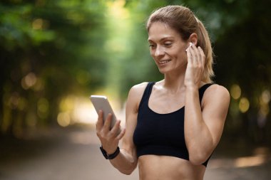 Pretty blonde middle aged athletic woman in black sportswear answering phone call while training outdoors at public park, pressing on earbud and looking at cell phone screen, copy space