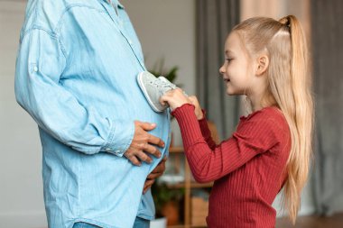 Loving little girl placing baby shoes at pregnant moms belly while playing and bonding, enjoying spending time together at home, side view