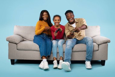 Family time activities. Overjoyed african american parents and their daughter playing video games, sitting on sofa over blue background. Dad and child holding joysticks