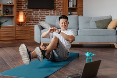 Happy asian middle aged man doing exercises for legs and abs on mat on floor in room interior and watching workout on computer. Online trainer, weight loss and body care