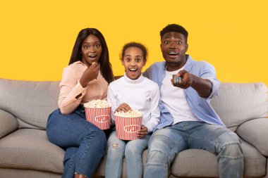 Shocked black parents and their daughter watching movie on TV, eating popcorn while sitting on sofa over yellow background. Family watching television