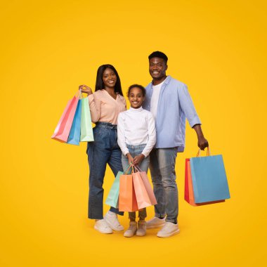 Seasonal sales concept. Happy african american family of three standing with shopping bags, excited black parents and their little daughter posing over yellow background
