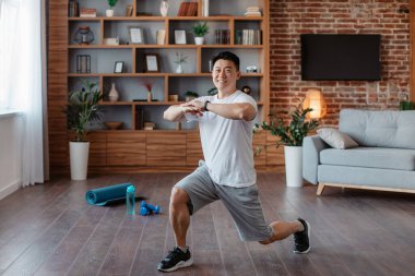 Athletic asian mature man making lunge, doing exercises, working out legs muscles at home and smiling at camera. Middle aged korean male practicing domestic sports