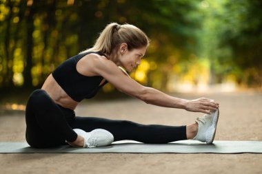 Sporty blonde woman stretching on yoga mat at public park, active middle aged lady having workout outdoors, enjoying morning activity, side view, copy space. Open air fitness concept