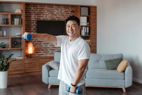 Home sports. Happy asian middle aged man doing fitness exercises with weights, using dumbbells and smiling. Excited male strengthening his arm muscles, leading healthy lifestyle