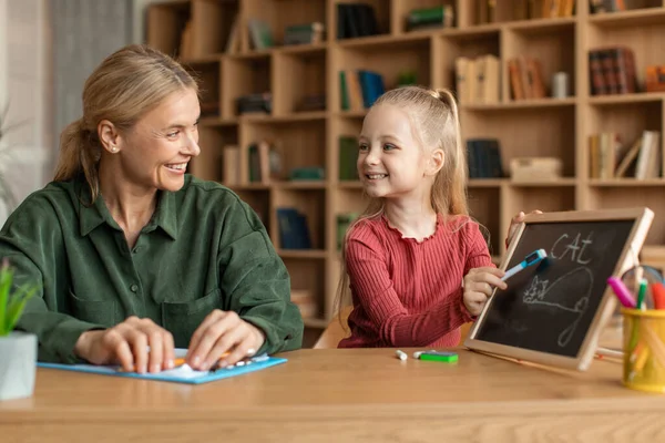 Female speech therapist teaching happy little girl alphabet, child writing word cat on blackboard with chalk. Speech-language therapy for children concept