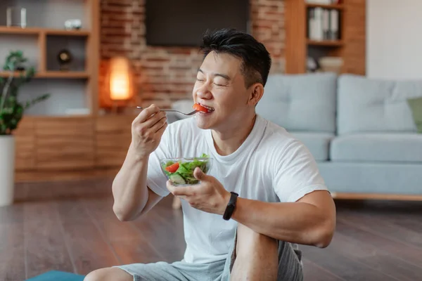 Fit asian middle aged man holding fork and bowl with fresh vegetable salad, sitting in living room interior, resting after home workout. Active mature male having balanced meal