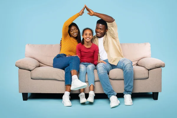 Family housing. Young black parents joining arms making symbolic roof above their daughter, sitting on sofa over blue background. Family dreaming about new house. Real estate concept