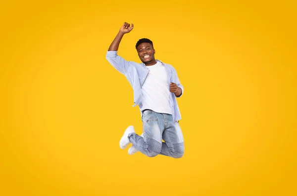 Carefree excited black man in casual outfit jumping in the air and smiling over yellow studio background, expressing positive emotions, full length shot, copy space