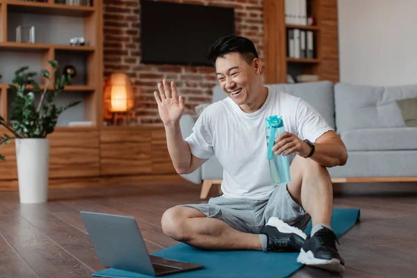 Online domestic training. Happy asian man waving to webcam, sitting on yoga mat in front of laptop, exercising online with coach in living room interior