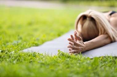 Closeup shot of unrecognizable blonde lady lying on yoga mat with hands crossed in public park, athletic woman having yoga practice outdoors in summer, panorama with copy space