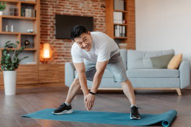 Active asian mature man doing exercises, stretching legs muscles at home on fitness mat, full length. Middle aged male practicing domestic sports, copy space