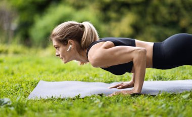 Motivated athletic blonde woman standing in cobra pose on fitness mat, sporty lady practicing yoga outdoors at public park, stretching her body, working on arms strength, side view, copy space