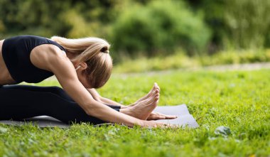 Side view of slim well-fit blonde lady sitting on fitness mat, reaching her knees with forehead, stretching her body in public park, doing yoga outdoors, panorama with copy space