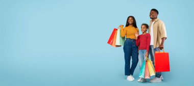 Family shopping concept. Happy african american parents and their daughter carrying colorful shopper bags over blue background, panorama with free space