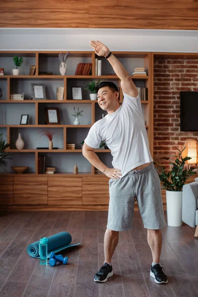 Active asian mature man in sportswear doing exercise and warm-up in living room interior. Sports, body care, weight loss and workout concept, crop