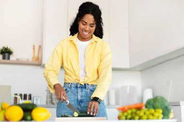 Dinner preparation. Happy black woman cutting cucumber, cooking vegetable salad, standing in kitchen interior. Housewife preparing fresh healthy meal
