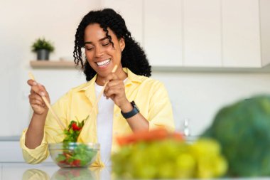 Happy black lady enjoying cooking, preparing healthy vegetable salad and mixing ingredients in bowl, standing in kitchen, copy space. Dieting and recipes concept