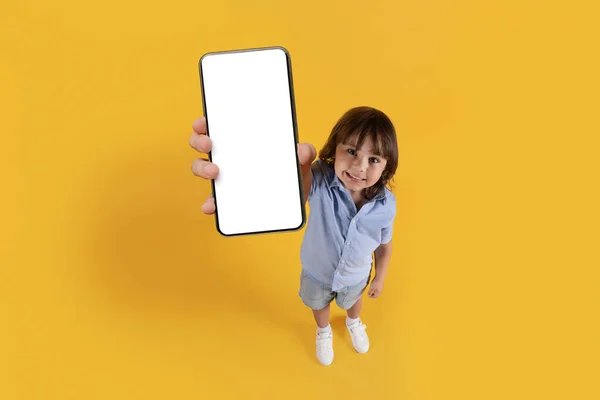 Mobile app for everyone. Cute excited little boy showing smartphone with white blank screen and smiling, above view portrait, free space