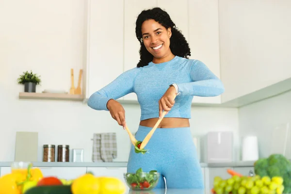 Making healthy meal. Happy fit african american lady mixing fresh vegetable salad in kitchen interior, cooking dinner and smiling at camera
