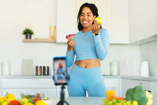 Choice of healthy or junk food. Fit black woman recording video on cellphone, showing donut and apple, shooting vlog in kitchen. Sporty lady talking about her snack