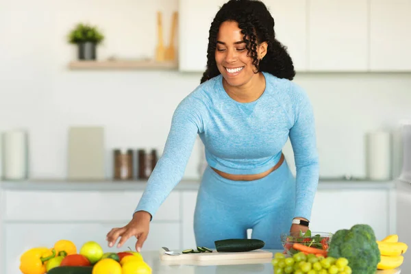 Happy black fit lady preparing fresh vegetable salad, cutting cucumber and taking tomatoes, standing in light modern kitchen interior and smiling