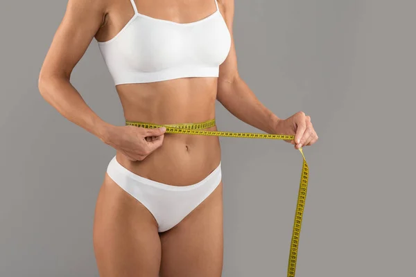 Weight Loss Concept. Closeup Shot Of Slim Young Woman In Underwear Measuring Waist With Yellow Tape, Unrecognizable Fit Female Checking Waistline Size While Posing Over Grey Background, Copy Space