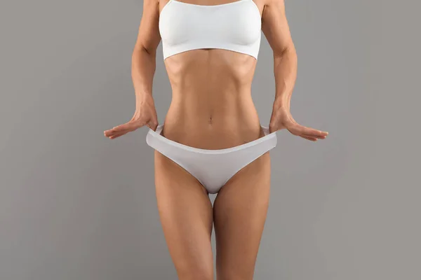 Unrecognizable fit woman with slim body in underwear pulling up white panties, young female with perfect sporty figure and flat abdomen standing isolated on grey studio background, copy space