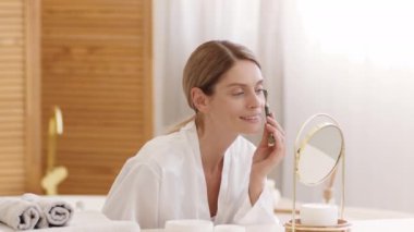Domestic spa procedures. Positive beautiful middle aged woman making facial skin lifting massage with green stone massager, looking at mirror at home, slow motion, free space