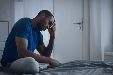 Unhappy African American Man Suffering From Depression Thinking About Problems And Feeling Lonely Sitting In Modern Bedroom At Home. Mental Health Issues. Side View, Low Angle