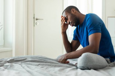 Male Depression. Black Guy Thinking About Issues Feeling Lonely And Unhappy Touching Head Sitting On Bed In Modern Bedroom At Home. Mental Health Problem. Side View