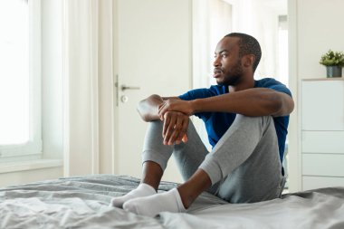 African American Man Thinking About Problems Looking Aside Having Depression Sitting On Bed In Modern Bedroom Indoors, Wearing Pajamas. Male Mental Health Concept