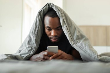 Sleepy African American Man Using Cellphone Texting Lying Covered With Blanket Lying In Bed In Modern Bedroom Indoor. Gadgets And Technology Concept. Selective Focus