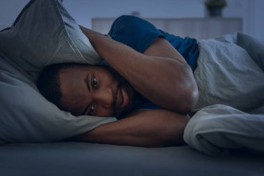 Black Man Having Insomnia Suffering From Anxiety Covering Ears With Pillow Posing Lying In Bed Looking At Camera In Modern Bedroom At Home. Sleeplessness Problem Concept. Low Light