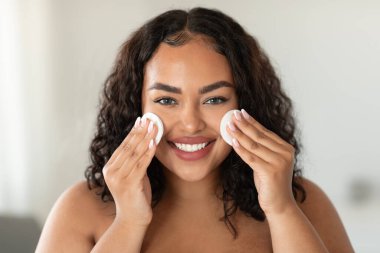 Facial skincare. Happy black chubby woman cleansing skin with cotton pads and smiling at camera, removing make up in bathroom, enjoying beauty routine, closeup headshot